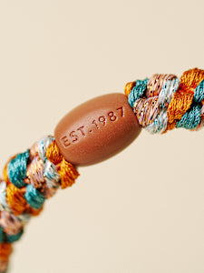 Close-up of a braided cord bracelet with orange, teal, and cream threads wrapping around a smooth brown oval bead engraved with 'EST. 1987' on a warm beige background.