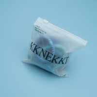A small translucent plastic pouch containing a pair of round silver hoop earrings, photographed on a smooth light blue background. The pouch has the brand name 'KKNKKEK' printed on it.
