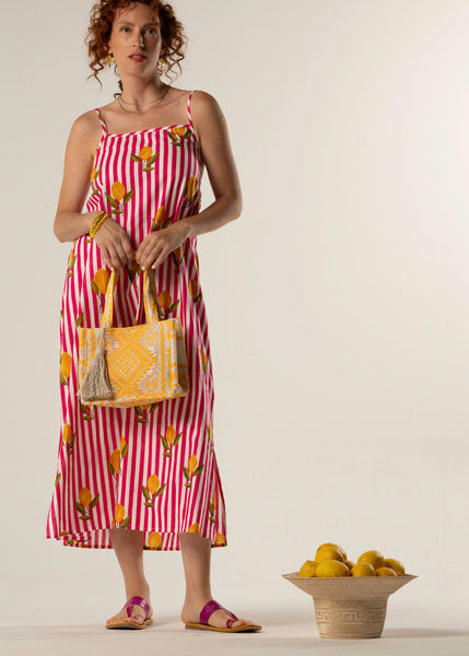 Woman in a red and white striped dress with fruit pattern holding a matching bag, standing next to a bowl of fruit on a plain background.
